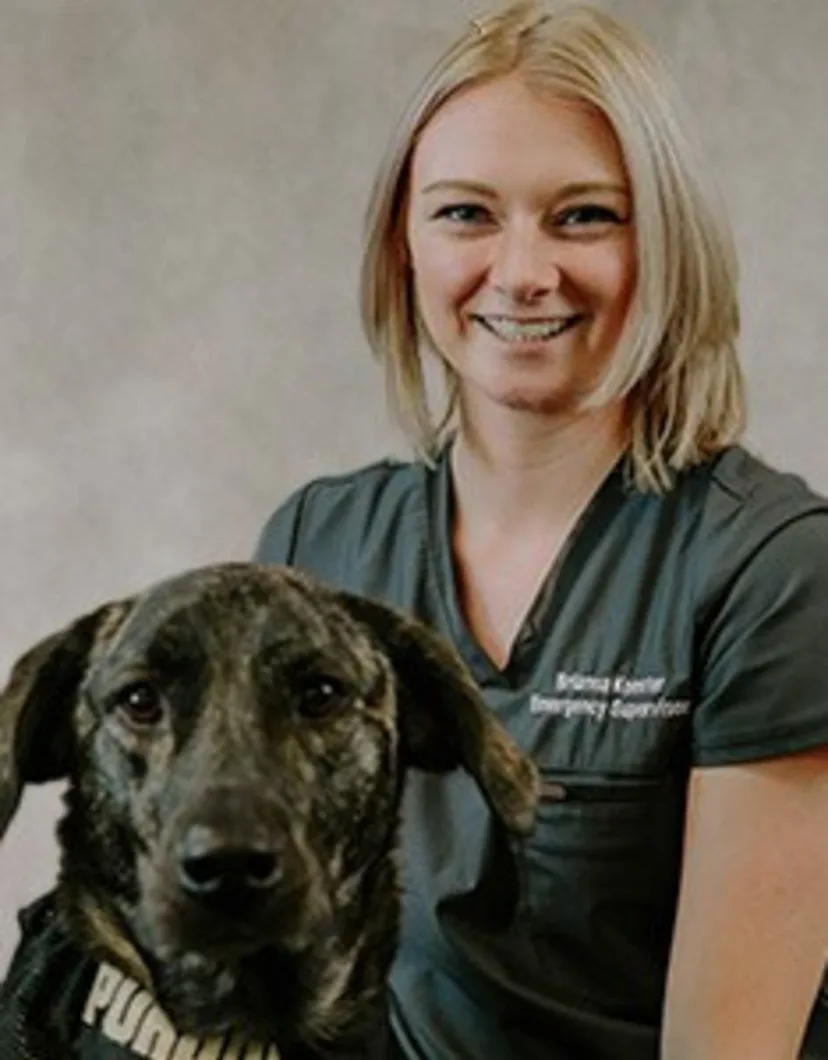 Brianna Koester smiling in front of a grey backdrop kneeling next to a large black and brown dog
